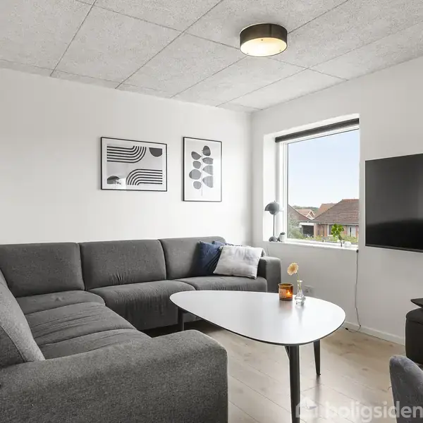 Grå hjørnesofa står i stue med lys trælaminatgulv. Et ovalt stuebord i hvidt står foran sofaen. Væggen har to abstrakte billeder. Vindue med udsigt. TV monteret til højre.