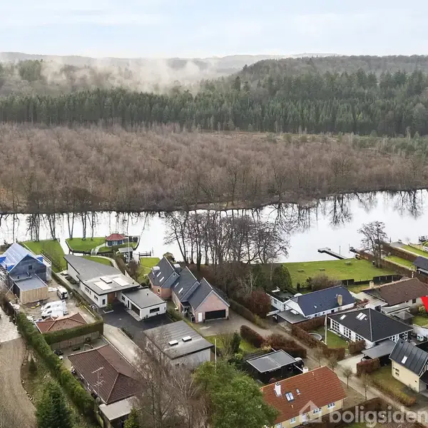 Et parcelhus er markeret med en rød pil tæt ved en flod. Huset ligger i en række af huse omgivet af en skovklædt bakkelandskab.