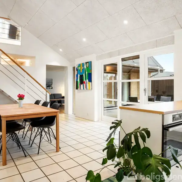 Et rummeligt spiseområde har et træbord med sorte stole. En trappe fører op til en øverste etage, mens grønne planter dekorerer rummet. Lyse fliser og et moderne køkken i baggrunden.