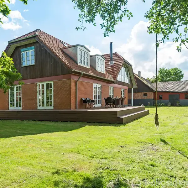 Et hus i mursten og træ står i en have med en stor græsplæne. En træterrasse med havemøbler ses ved huset, og et gyngereb hænger fra et træ.