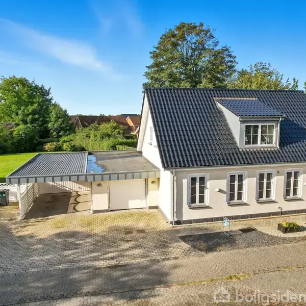 Hvidt hus med sort tag, står ved en flisebelagt gårdsplads, omgivet af grønne træer og en velplejet græsplæne. Solrigt vejr og klar blå himmel i baggrunden.