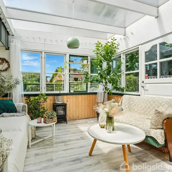 Lys indendørs veranda med to sofaer, et rundt bord med blomster, og flere planter. Store vinduer lukker lys ind og udsigt til en have.