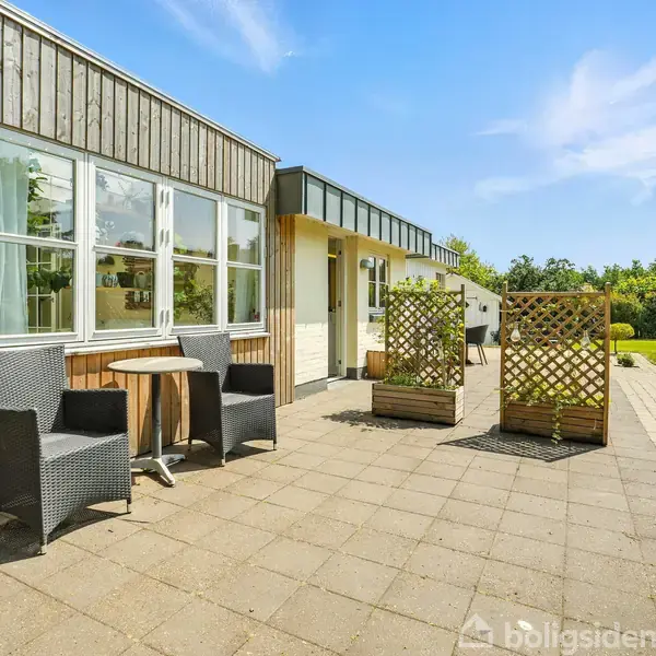 Hus med træbeklædning står på flisebelagt terrasse med havemøbler. To flettede afskærmninger adskiller områder. Græsplæne og buske i baggrunden. Klar blå himmel og sollys.