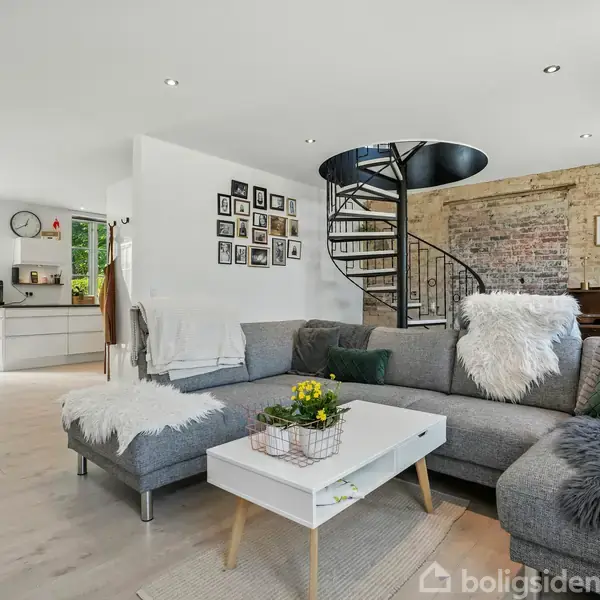 Gråt hjørnesofa er centralt placeret i en lys stue med en hvid bord, spiraltrappe i baggrunden og åben adgang til køkkenet med lyse træmøbler og store vinduer.