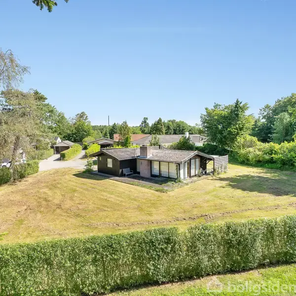 Et hus står stille på en stor, åben græsplæne omgivet af hække og træer under en klar, blå himmel i et stille boligområde.