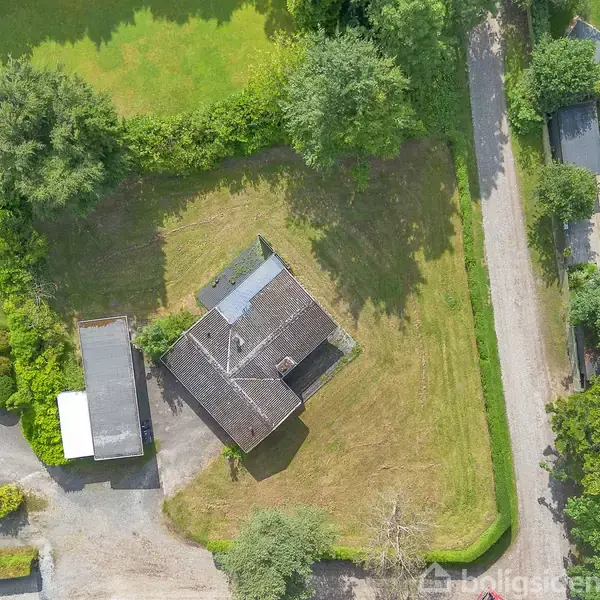 Et hus med gråt tag står midt i en græsplæne omgivet af træer og buske. En grusvej løber langs den ene side, og der er en tilstødende bygning.