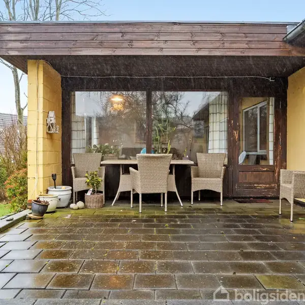 En terrasse med havemøbler står under et halvtag. Bord og stole er placeret på en flisebelagt overflade. Der er planter og en græsplæne til venstre.