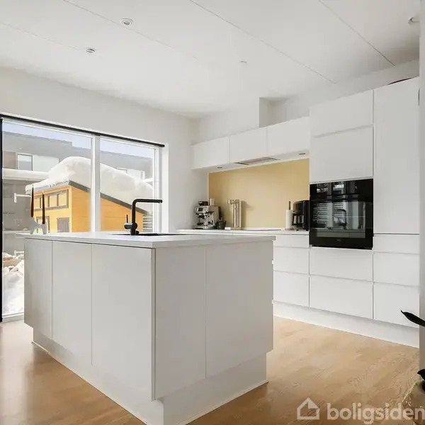 Køkken med hvidt køkkenø og sort vandhane, set i dagslys gennem store vinduer. Minimalistisk indretning med hvide skabe og indbygget ovn, omgivet af lyst trægulv og snedækkede udendørs omgivelser.