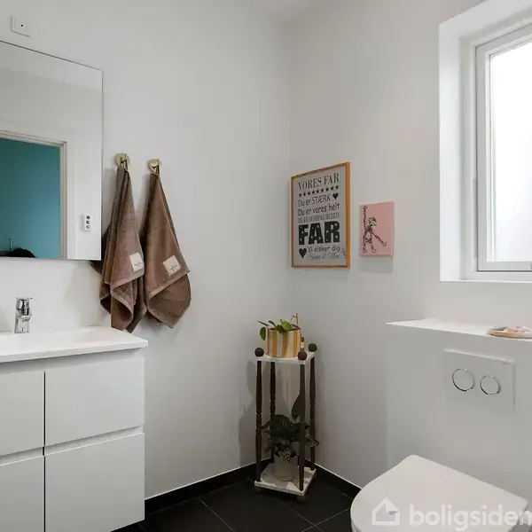 Hvidt badeværelse med en moderne vask og spejl. To håndklæder hænger på kroge. På væggen er der et skilt med teksten: "FAIR". En lille plante står ved vinduet.