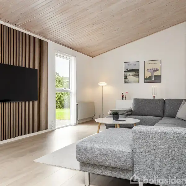 Grå sofa står i en stue med trægulv og et skrånede træloft. Et TV hænger på en væg med træpaneler. Vinduet viser en have. To plakater hænger over sofaen.