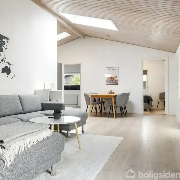 Grå sofa med tæppe på i moderne stue; kort over verden på væggen. Lys træloft med loftvinduer. Rundt sofabord, spisebord med stole bagved. Lyse trægulve, lyse rum.