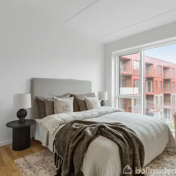 En seng med puder og en plaid ligger opredt i et lyst, minimalistisk soveværelse. En natbordslampe står ved siden af, og et vindue viser røde bygninger udenfor.