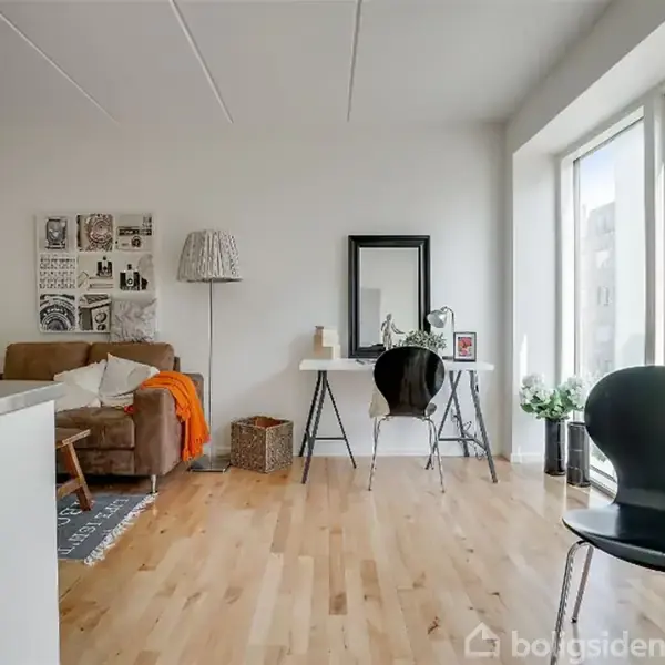 Stue med trægulv, en brun sofa til venstre, en bordlampe og et lille sort bord med spejl til højre, store vinduer bagest. Ingen tekst fundet.