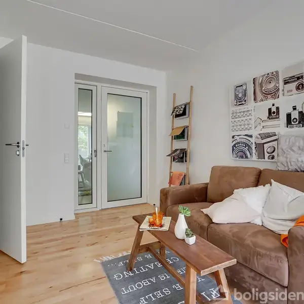 Brun sofa står i lys stue, dekoreret med puder og tæppe. Ved siden af er et lavt bord med drikkevarer. Væg med kunstværker til højre, åben dør til venstre.