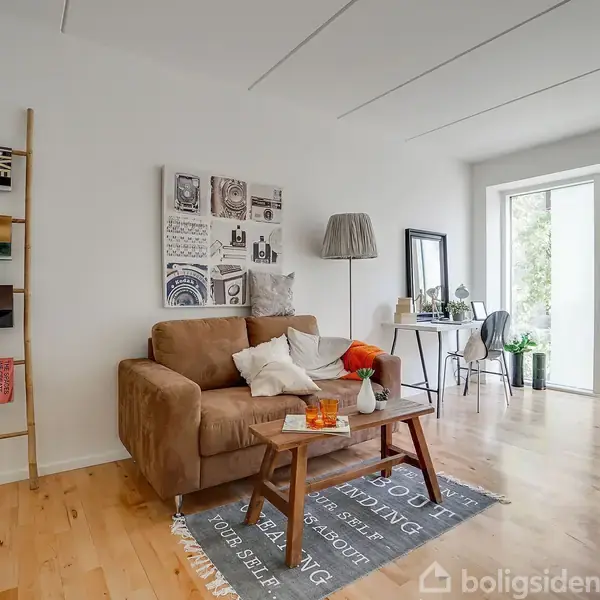 Brun sofa står stille ved sofabord, dekoreret med blomster og stearinlys, i en lys stue. Stige med bøger og plakater pryder væggen. Store vinduer giver udsigt til natur.