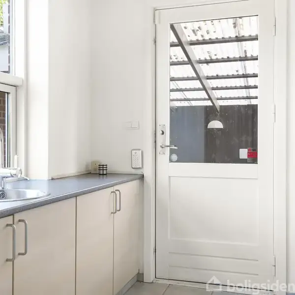 Hvid dør med glasrude åbner ud til overdækket terrasse; køkkenbord langs væg med vask og skabe; lysindfald fra vindue; ren, minimalistisk indretning.