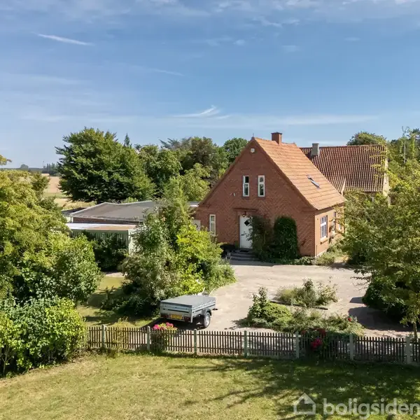 Et rødt murstenshus står omgivet af frodige træer og buske. Foran huset er der en indkørsel med en trailer. Området er åbent med en blå, skyfri himmel.