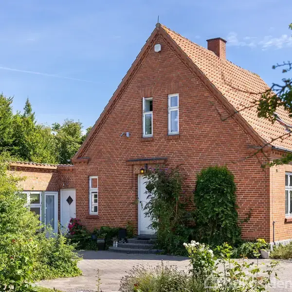 Et rødt murstenshus med tegltag står stille i en have med blomster og buske. Sollys skinner på huset, omgivet af grønne træer og klar blå himmel.