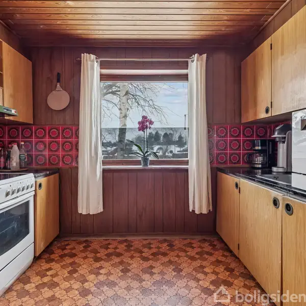 Et køkken med træpaneler har en ovn til venstre, hylder med krydderier, og til højre skabe og en mikrobølgeovn. En orkidé står i vindueskarmen med udsigt til have.