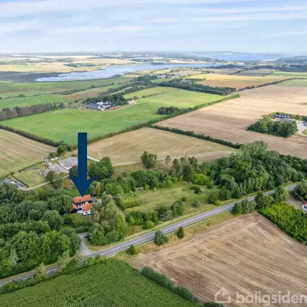 Hus markeret med blå pil står blandt træer. Omgivet af marker og veje i et landligt område. En sø kan ses i baggrunden.