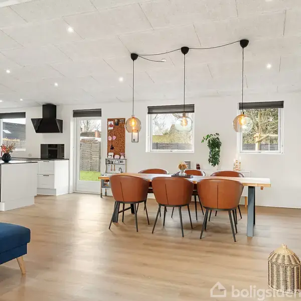 Et åbent køkken-alrum med træbord omgivet af skindstole i brun læder. Tre hængelamper svæver over bordet. Store vinduer giver udsigt til en have.