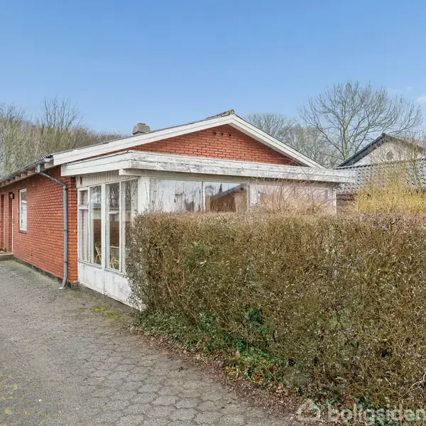 Rødt murstenshus med stor glasveranda står stille langs en flisebelagt indkørsel. Omkring huset er en velholdt hæk og træer i baggrunden. Himlen er klar og blå.