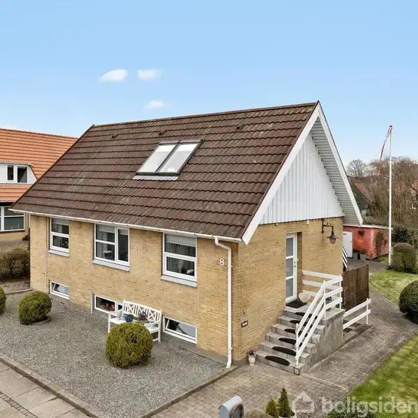 Et gult murstenshus med brunt tegltag har skrånende tag og ovenlysvinduer. Huset er omgivet af en grusbelagt gårdsplads og velplejede buske, med en flagstang i haven. Nummer 8 ses på huset.