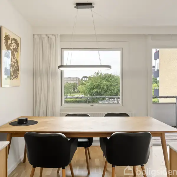Et spisebord af træ er omgivet af tre sorte stole og en moderne lampe over det. Bordet står i et lyst rum med hvide vægge, en vindueudsigt til grønne træer og en balkon.