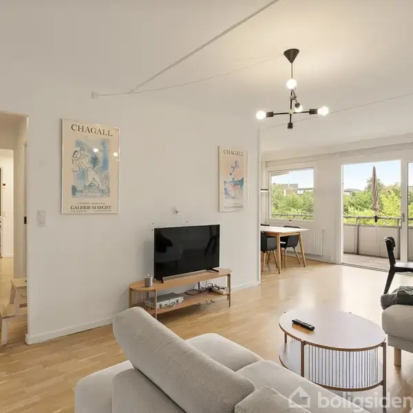 Stue med beige sofa, fladskærms-tv på væggen, to Chagall-plakater, og stort vinduesparti med udsigt over grønne træer. Rundt bord ved sofaen, spisebord i baggrunden.