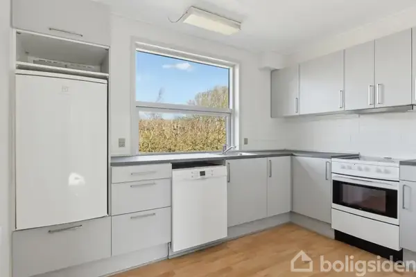 Køkken med hvide køkkenskabe og lysegrå bordplader, hvor opvaskemaskine og ovn indbygget i skabene. Vindue med udsigt til grøn busk omkring huset.