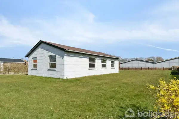 Et enkelt, hvidt hus med skråt tag står stille på en grøn græsplæne. Omkring huset ses en lav hæk og andre boliger i baggrunden under en blå himmel.