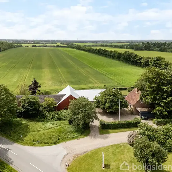 Et landsted ligger tæt på en vej, omgivet af frodige, grønne marker og tætte skovområder under en klar, blå himmel.
