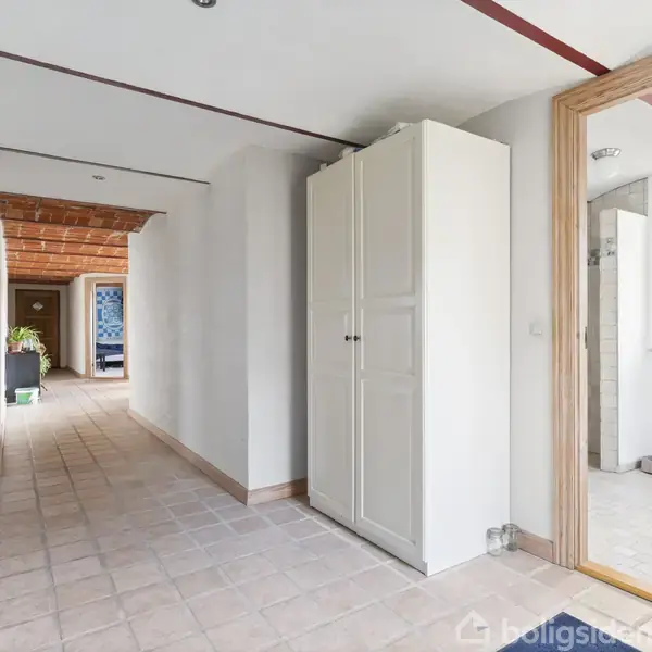 Et skab står stille i en lys gang med flisegulv, der fører til åbne døre. Rummet har hvidmalede vægge og trædørkarme med et vindue ved siden af.