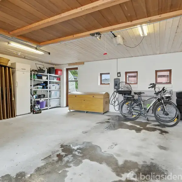 Garage med to cykler ved siden af hinanden, placeret mod en hvid væg med dækstable og opbevaringshylder til venstre. Rummet har et træloft med lysarmaturer.