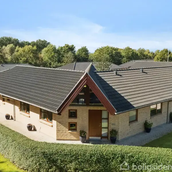 Hus med vinkeltag står stille omgivet af en velplejet have med tætklippede hække og græsplæne. Baggrunden består af høje træer under en klar blå himmel.