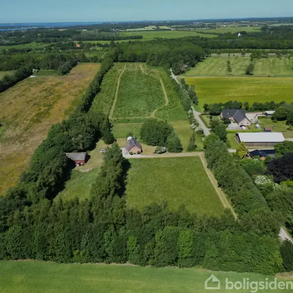 En gård omgivet af marker og grønne områder. Bygningerne inkluderer et hus og en lade. Over konteksten spredes landskabet med skov og marker. Flask grønne områder dominerer scenen.