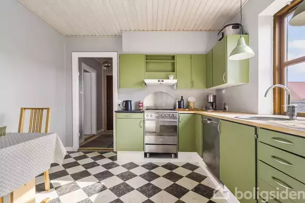 Et køkken med grønlige skabe og en rustfrit stål ovn viser en simpel og funktionel indretning. Gulvet har et sort-hvidt ternet mønster, og der er en træstol ved et bord med en hvid dug.