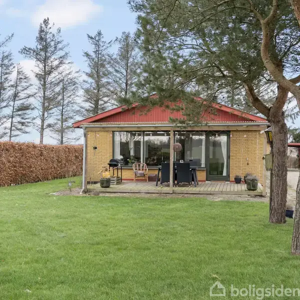 Et gult murstenshus med rødt tag står på en grøn græsplæne. Huset har en terrasse med bord og stole foran. Omkring huset ses træer og en hæk.