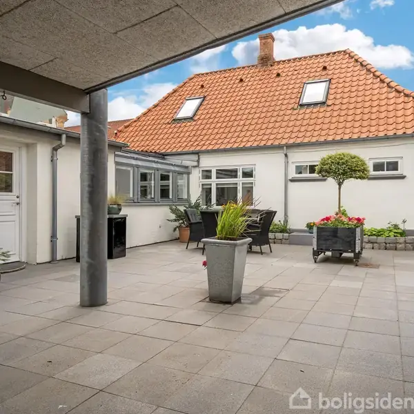 En flisebelagt gårdhave med flere plantekrukker står mellem to huse med rødt tegltag og grå vinduer. Der er stakit til højre og blå himmel med skyer.