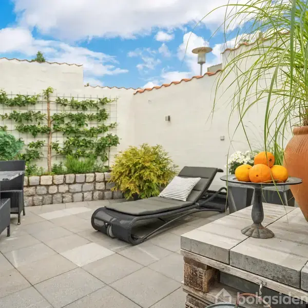 Liggestol ligger på terrasse med fliser, omkranset af hvidmalet mur med planter. Et bord med appelsiner står ved siden af en stor krukke og lanterne.