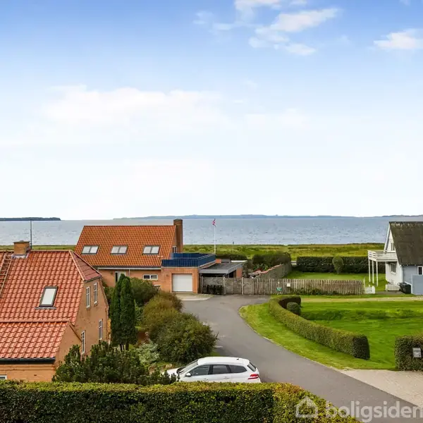 Røde og hvide huse står ved en vej nær grønne hække, med havudsigt og blå himmel i baggrunden.