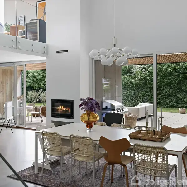 Spisebord omgivet af stole står i en moderne spisestue med en stor glasdør, der åbner til en terrasse med planter. I baggrunden er der en tændt indendørs pejs.