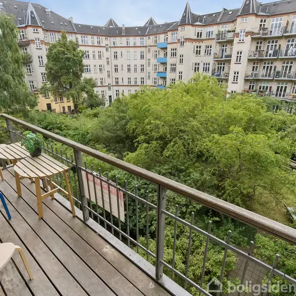 Balkon med træmøbler og en lille grill står stille. Den vender ud mod en stor grøn gårdhave omgivet af høje, ældre lejlighedsbygninger med mange vinduer.