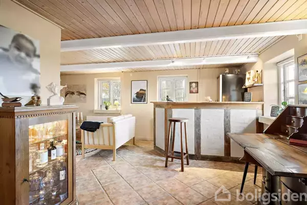 Et stueområde indeholder en sofa og en bar med barstole. Møblerne er omgivet af træpaneler, kunst på væggene og en vitrine med flasker til venstre.