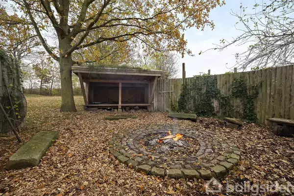 Et lille bål brænder i en cirkel af sten. Foran et shelter i skoven, omgivet af faldne blade og et hegn beklædt med vinranker.