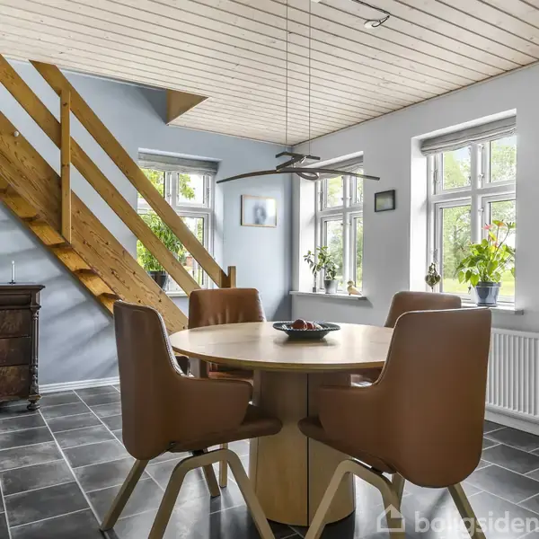 Et rundt bord med fire læderstole står i et rum med mørke fliser. Trappen i træ fører opad. Vinduerne slipper lys ind, og der er planter og billeder på væggen.