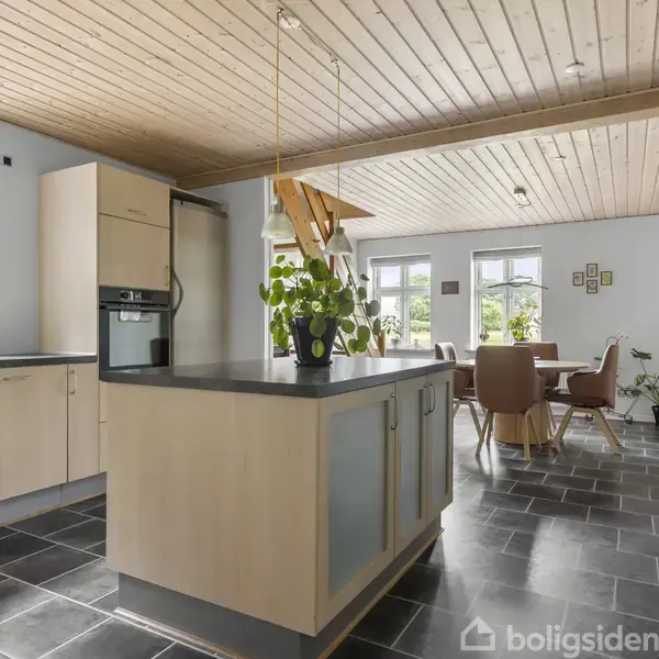 Køkken med træloft og sorte fliser, centralt placeret køkkenø med en plante. I baggrunden er der et spiseområde med rundt bord og brune stole ved store vinduer.