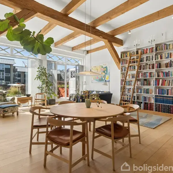 Et rundt spisebord med fire stole står centralt under en moderne lampe. Rummet har store vinduer, træbjælker, bogreoler langs væggen og grønne planter, der skaber en lys atmosfære.
