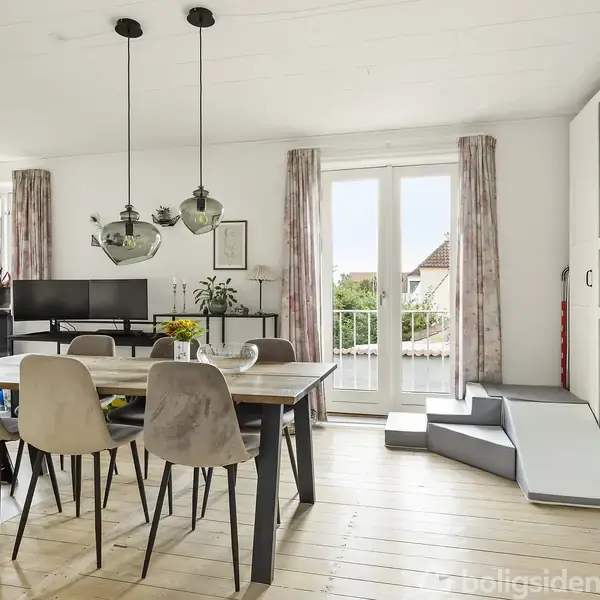 Spisebord med seks stole står i en lys stue med lyse trægulve. To lamper hænger over bordet. Franske døre åbner ud til en balkon med udsigt til bygninger.