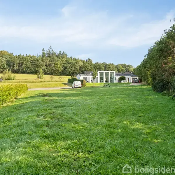 En grøn plæne strækker sig mellem to moderne huse på landet, omgivet af træer i baggrunden under en lys himmel. En bil er parkeret ved huset i midten.